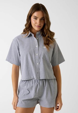 Junge Frau mit langem welligem Haar, die ein kurzärmeliges, kariertes Hemd mit Knöpfen und passende Shorts mit elastischem Bund vor einem einfarbigen Hintergrund trägt.