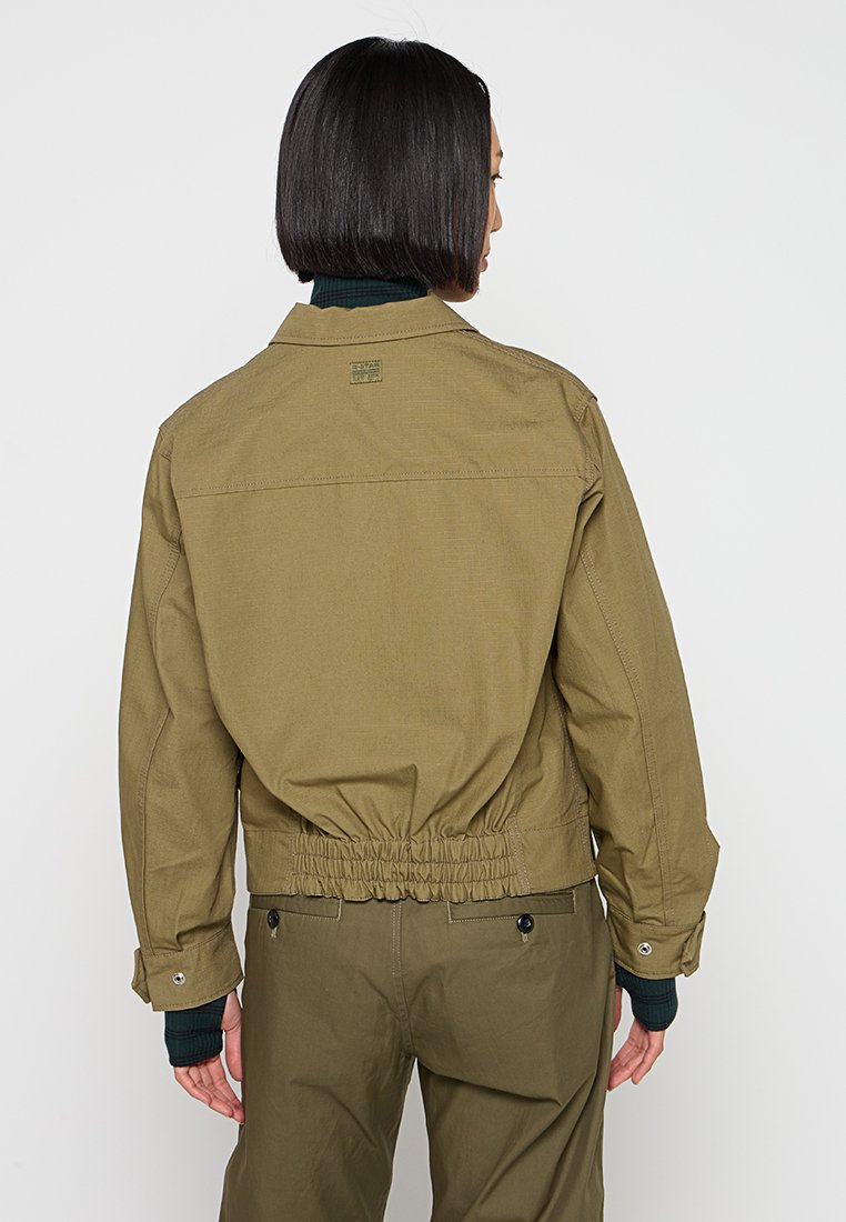 Olivenfarbige Jacke mit lockerer Passform, elastischem Bund und Druckknopfmanschetten. Verfügt über ein Etikett auf der Rückseite und strukturierte Schultern.