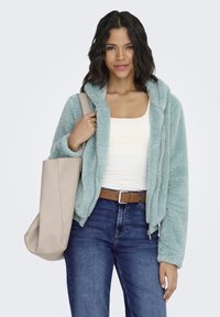 Flauschige türkisfarbene Kapuzenjacke mit Reißverschluss vorne, getragen über einem weißen Hemd, kombiniert mit dunklen Jeans und einer beigen Tragetasche.