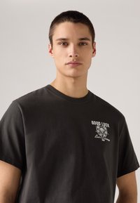 Black cotton t-shirt featuring “GOOD LUCK” printed in white with decorative graphics on the front. Crew neckline and short sleeves.