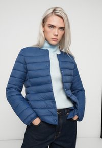 Modrá puffová bunda s horizontálne prešívaným dizajnom, so cvočkami a okrúhlym výstrihom, v kombinácii so svetlomodrým rolákom a tmavými džínsami.
