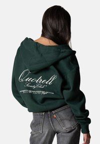 Dunkelgrüner Kapuzenpullover mit weißem Logo-Schriftzug auf der Rückseite. Aus Baumwollmaterial, lockere Passform, mit gerippten Bündchen und Saum.