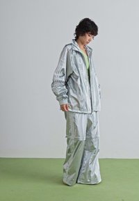Persoon in een losse, glanzende zilveren jas en wijde broek die op een groene vloer staat tegen een eenvoudige lichtgrijze muur.
