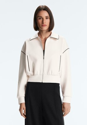 Frau mit glattem braunem Haar, die eine weiße Jacke mit Reißverschluss und schwarzer Borte sowie schwarze Hosen vor einem einfarbigen Hintergrund trägt.