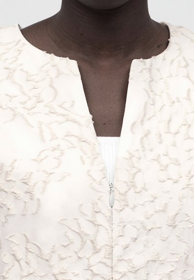 Κοντινό πλάνο ενός σκουρόχρωμου ατόμου που φοράει υφασμάτινη εκρού ζακέτα με κεντρικό φερμουάρ, μερικώς ανοιχτό πάνω από λευκό τοπ.