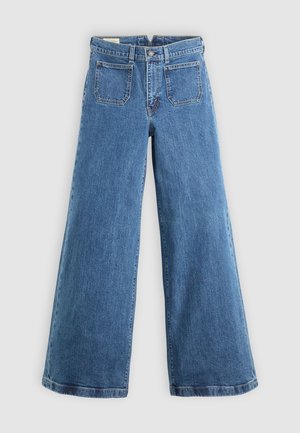Jean blue à taille haute et jambe large avec poches avant à rabat, passants de ceinture, fermeture à bouton et braguette zippée sur fond uni.