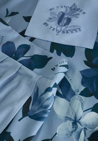 Šviesiai mėlyna medžiaga su tamsiai mėlynu gėlių raštu ir siuvinėta etikete, ant kurios užrašyta "b by Ted Baker" su stilizuotu „b“ ir lapais.