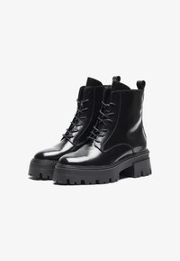 Bottines noires en cuir avec une finition brillante, à lacets et avec des semelles épaisses et robustes. Équipées de languettes pour un enfilage facile.