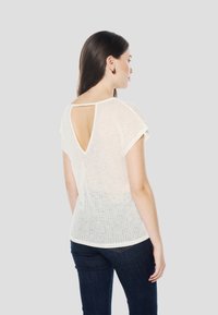 Krēmkrāsas īsām piedurknēm tops ar atvērtu aizmuguri, caurspīdīgu tekstūru un apaļu apakšu. Kopā ar tumši zilām džinsām.