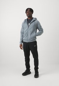 Gray zip-up hoodie with front pockets, worn over a navy T-shirt, paired with black sweatpants featuring a white logo, and black sneakers.