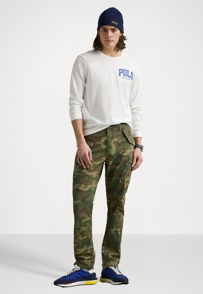 Λευκό πουκάμισο με μακριά μανίκια που γράφει "POLO" σε μπλε, καμουφλάζ cargo παντελόνια και μπλε αθλητικά παπούτσια με κίτρινες λεπτομέρειες. Ένα κ�άπ της ναυτικού χρώματος ολοκληρώνει την εμφάνιση.