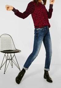 Röd och svart mönstrad långärmad topp i kombination med blå denimjeans och svarta ankelskor. Vit metallstol i bakgrunden.