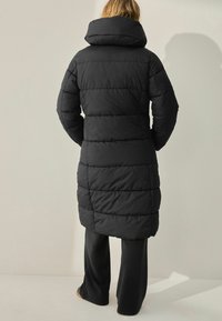 Une personne se tient de dos devant la caméra, portant un long manteau d'hiver noir et un pantalon noir, sur un fond neutre.