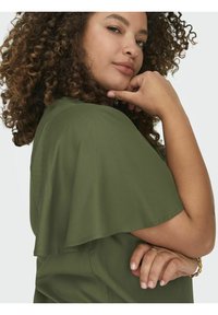 Olivinės spalvos blusa su plazdančiais rankovėmis, minkšta tekstūra ir laisvas siluetas. Modelis turi garbanotus plaukus, poilsiauja su žandikauliu ant rankos, demonstruodama subtilius papuošalus.