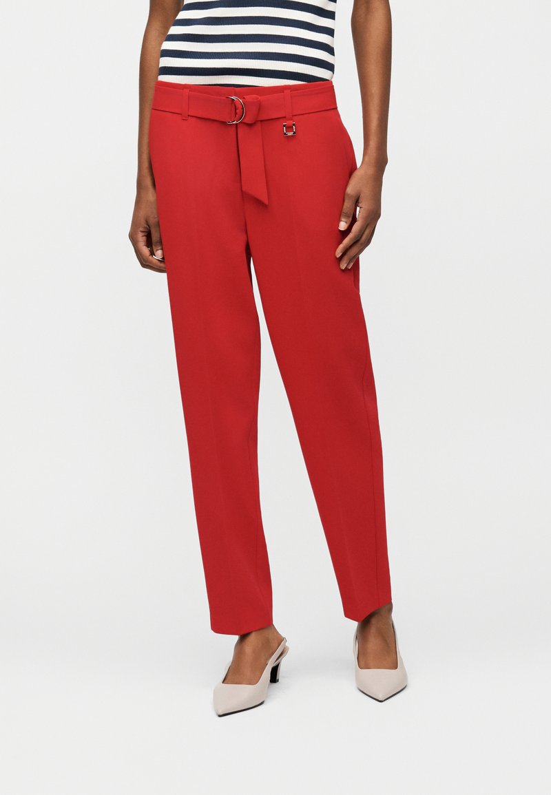 Femme portant un pantalon taille haute rouge avec une ceinture en tissu assortie et des talons blancs à bout pointu, assortis à un haut rayé bleu marine et blanc.