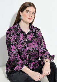 Camicia nera con bottoni, caratterizzata da un motivo floreale in tonalità di viola e teal, con maniche lunghe e una vestibilità comoda. Il modello è seduto contro uno sfondo bianco.