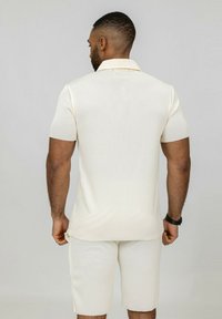 Polo à manches courtes couleur crème avec texture côtelée, associé à un short côtelé assorti. Col et fentes sur les côtés pour faciliter les mouvements.