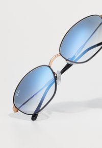 Ray-Ban päikeseprillid suurte siniste gradient-läätsedega, must metallraam, kuldsed detailid ja läbipaistvad nina padjad. Nurgeline disain modernse välimuse jaoks.