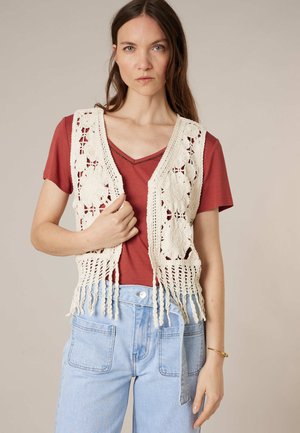 Femme portant une chemise à manches courtes couleur rouille, un gilet en crochet crème avec des motifs floraux, et un jean taille haute bleu clair.
