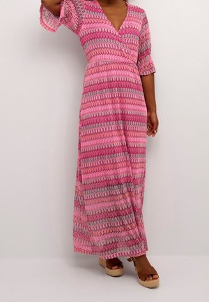 Kobieta ubrana w długą sukienkę maxi z wzorem w odcieniach różu i fioletu, z rękawami do łokcia, oraz brązowe sandały na koturnie.