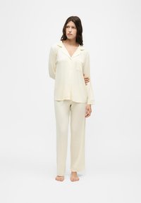 Femme debout pieds nus, portant un haut de pyjama ample crème à manches longues avec col et un pantalon large assorti.