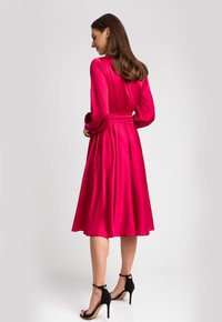Fuksjowa satynowa sukienka z długimi rękawami i dopasowaną talią. Rozkloszowana spódnica sięgająca poniżej kolan, w połączeniu z czarnymi sandałami na wysokim obcasie z paskami.