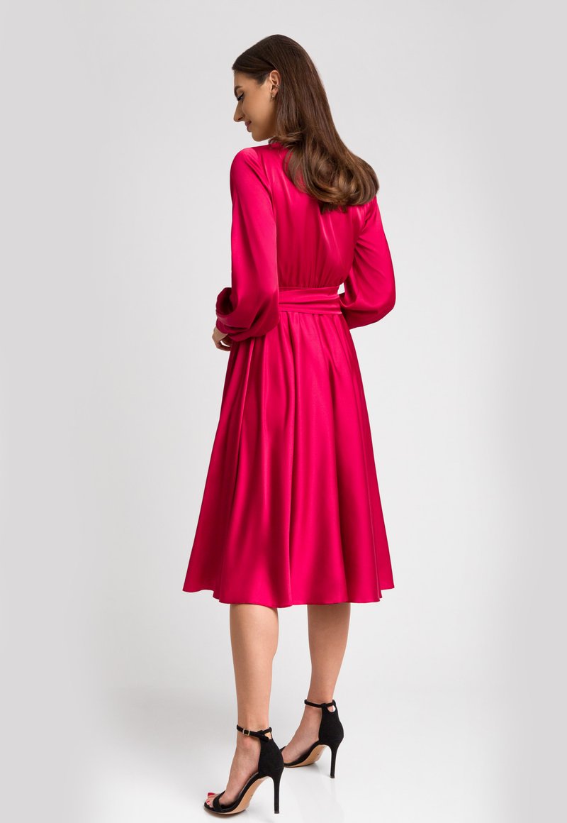 Fuksjowa satynowa sukienka z długimi rękawami i dopasowaną talią. Rozkloszowana spódnica sięgająca poniżej kolan, w połączeniu z czarnymi sandałami na wysokim obcasie z paskami.
