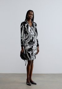 Svart-hvitt marmormønstret omslagskjole med lange ermer, definert midje og midtlårlengde, kombinert med en liten svart håndveske.