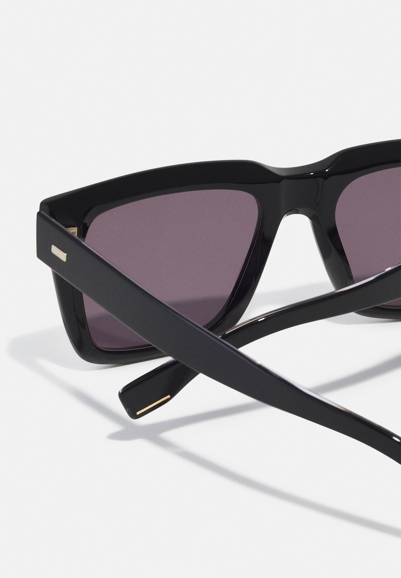 Lunettes de soleil noires avec un design géométrique, dotées de verres gris et d'accents dorés sur les branches. La monture est brillante avec une construction épaisse.