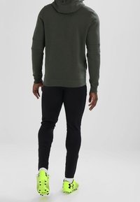 Sudadera capucha verde con un acabado texturizado, combinada con pantalones ajustados negros y zapatillas deportivas amarillas brillantes con tachuelas.