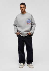 Grijze oversized sweatshirt met een voorkantzak waarin blauwe en rode tekst staat. Gecombineerd met donkere denim jeans en sneakers in neutrale tinten.