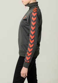 Grå atletisk jacka med hög krage, med ett djärvt röd- och grått chevronmönster på ärmarna. Slät, lättviktsstoff.