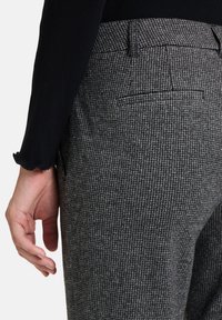 Spodnie w szary wzór houndstooth o dopasowanym kroju, z tylną kieszenią i delikatnie teksturowanym materiałem. Widoczna czarna bluzka z długim rękawem.