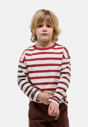Enfant blond aux cheveux mi-longs portant un t-shirt à manches longues rayé blanc et rouge et un pantalon foncé, ajustant une manche.
