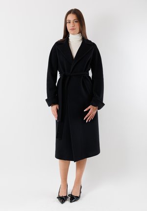 Cappotto di lana nero con vita cinturata, maniche lunghe e collo a scialle. La modella indossa un dolcevita crema e decolté nere a punta.