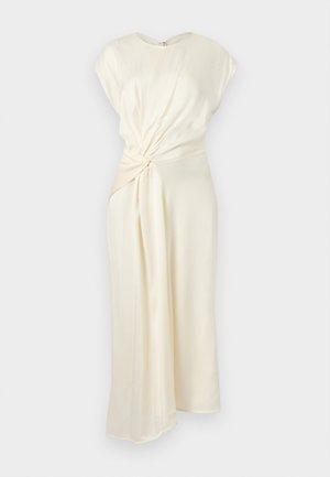 Moteris tamsiais plaukais, vilkinti kreminės spalvos asimetrišką midi suknelę su mazgo detalėmis, juodus batelius su dirželiais, stovinti baltame fone.