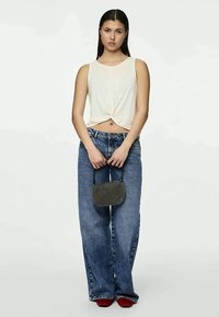 Junge Frau trägt ein ärmelloses weißes Crop-Top, weite blaue Jeans, rote Schuhe und hält mit beiden Händen eine kleine schwarze Handtasche.
