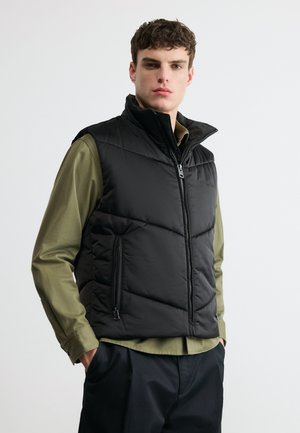 Joven con un chaleco negro acolchado sobre una camisa de manga larga verde oliva, de pie con las manos en los bolsillos contra un fondo blanco liso.