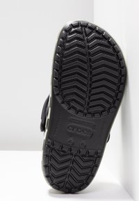 Semelle de chaussure Crocs noire avec des motifs de bande de roulement profonds, des marquages de taille et le nom de la marque embossés sur une surface blanche.