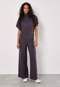 Lila Strick-Loungewear-Set mit einem kurzärmeligen Oberteil mit hohem Kragen und weit geschnittenen Hosen. Weicher Stoff. Kombiniert mit weißen Sneakers.