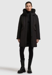 Sort vandtæt parka med hætte, frontlommer og lynlåse, med en mellemlang design og et glat, struktureret stof. Sorte leggings og støvler.
