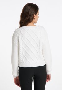 Weißer Strickpullover mit gerippten Bündchen und Saum. Die Rückseite zeigt strukturiertes Diamant- und Zopfmuster. Getragen mit schwarzen Hosen.