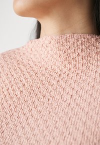 Pfirsichfarbendes Oberteil mit einem strukturierten, zerknitterten Design. Hoher Kragen mit einem unregelmäßigen Muster, aus einem leichten, weichen Stoff gefertigt.