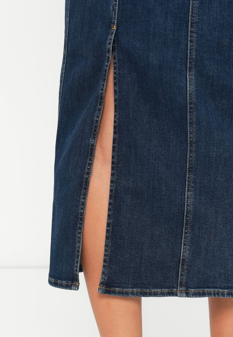 Benen gedeeltelijk zichtbaar door een zijsplit in een donkerblauwe denim rok met oranje stiksels, tegen een effen witte achtergrond.