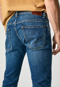 Denim jeans in a dark blue shade, featuring a textured surface, visible stitching, and distinctive back pockets with a V-shaped design.