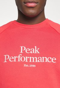 Rode sweatshirt met witte geborduurde tekst "Peak Performance" en "Est. 1986", met een ronde hals en raglan mouwen.