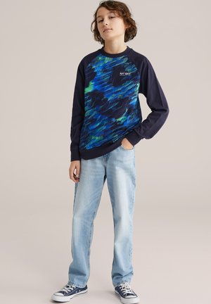 Jeune personne debout, la main dans la poche, portant un sweat-shirt bleu à motifs, un jean clair et des baskets bleu marine sur un fond uni.