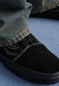 Black suede sneaker with white stitching, flat laces, and a rubber sole, partially obscured by light blue carpet and denim pants.