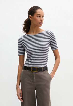 Frau steht mit einer Hand in der Tasche, trägt ein grau-weiß gestreiftes kurzärmeliges Oberteil, braune Hose und einen schwarzen Gürtel mit goldener Schnalle.