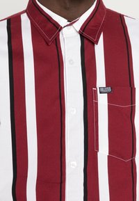 Rot-weißer gestreifter Hemd mit Knöpfen, schwarzen Akzenten, Kragen und einer Brusttasche mit einem "Hollister"-Label. Aus Baumwollmaterial.